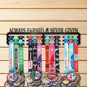 May include: Black metal medal display rack with the text "ALWAYS EARNED NEVER GIVEN". The rack holds several medals with colorful ribbons. The medals have various designs and logos, hanging against a wooden background.