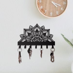 May include: Black metal key holder with a decorative mandala design. The key holder has six hooks and is mounted on a white wall. Several keys hang from the hooks. A wooden clock is visible in the background.