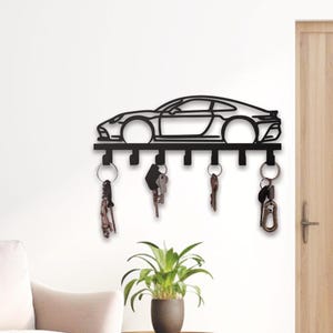 Può includere: Portachiavi in metallo nero a forma di auto sportiva. Il portachiavi ha sei ganci sotto la silhouette dell'auto, con diversi mazzi di chiavi appesi. Il portachiavi è montato su una parete bianca.