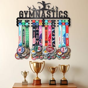 以下が含まれることがあります： 「GYMNASTICS」の文字と体操選手のシルエットが入った黒い金属製の体操メダルホルダー。下には、多数のメダルを保持するカラフルなリボンがあります。4つの金色のトロフィーが木製の表面に置かれています。