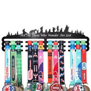 May include: Black metal medal display with the quote "Not All Those Who Wander Are Lost." The display holds several medals with colorful ribbons. The ribbons have text and designs. The top of the display has a silhouette of a scene with figures and trees.