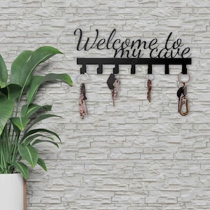 May include: Black metal key holder with the text "Welcome to my cave" in a cursive font. The key holder has six hooks, each holding a set of keys. The key holder is mounted on a white brick wall, with a potted plant in the corner.