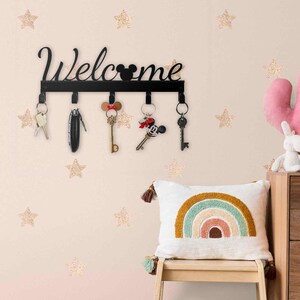 Könnte beinhalten: Schwarzer Metall-Schlüsselhalter mit dem Wort "Welcome" in Schreibschrift, mit einer Mickey Mouse Silhouette. Der Schlüsselhalter hat fünf Haken mit Schlüsseln daran. Die Wand ist mit goldenen Glitzer-Stern-Aufklebern dekoriert.