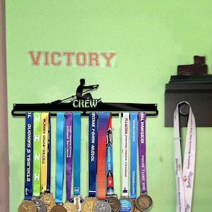 Puede incluir: Un estante de exhibición de medallas de metal negro con la palabra "CREW" y una silueta de un remero. El estante contiene varias medallas con cintas de colores. La palabra "VICTORY" está encima del estante.