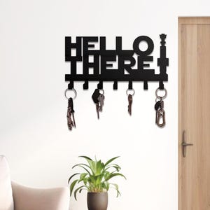 Pode incluir: Porta-chaves de metal preto com as palavras "HELLO THERE" recortadas. O porta-chaves tem vários ganchos com chaves penduradas. Uma pequena planta em vaso está por baixo.