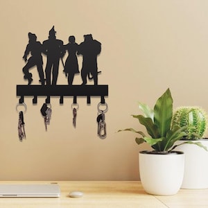 Puede incluir: Llavero de metal negro con siluetas de personajes de El Mago de Oz. El llavero tiene seis ganchos con llaves colgando. Está montado en una pared beige, con plantas en macetas en primer plano.