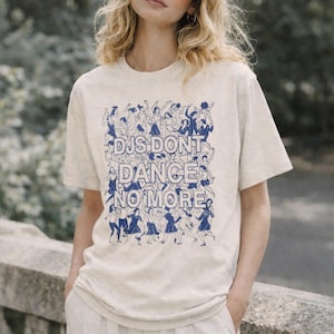 May include: A cream-coloured t-shirt featuring the blue text "DJs Don't Dance No More" and a graphic of dancing figures. The t-shirt is worn by a person wearing sunglasses.