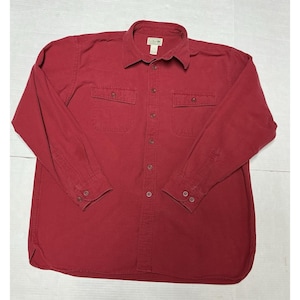 Puede incluir: Una camisa abotonada de manga larga en un tono rojo intenso. La camisa presenta dos bolsillos en el pecho, cuello y puños abotonados. La etiqueta de la marca es visible en la parte superior del pecho. La camisa está hecha de una tela resistente.