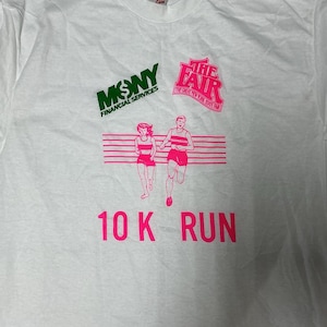 Puede incluir: Camiseta blanca con un gráfico rosa y verde. El gráfico incluye el texto "MSNY FINANCIAL SERVICES" en verde, "THE FAIR THE GREAT NEW YORK STATE FAIR" en rosa y una ilustración rosa de dos corredores. El texto "10 K RUN" también es rosa.