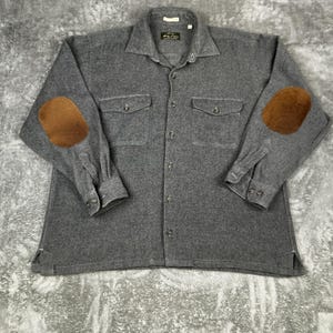 May include: A gray button-down shirt with two chest pockets and brown elbow patches. The long-sleeved shirt has a collared neckline and buttoned cuffs. The shirt is made of a soft, textured fabric.