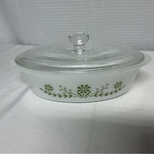Puede incluir: Fuente ovalada blanca con tapa de cristal transparente. El plato presenta un estampado floral verde en los laterales. La tapa tiene un pequeño asa redonda. Un clásico de la cocina.