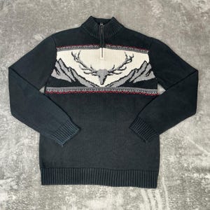 Puede incluir: Suéter de punto negro con cuello alto y cremallera de cuarto. El suéter presenta un panel blanco con un diseño de montaña y cabeza de ciervo, acentuado con detalles rojos y grises. Puños y dobladillo acanalados.
