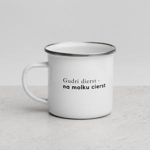 May include: White enamel mug with a silver rim and handle. The mug features the text "Gudri dierst - na molku cierst" in black, centered on the front. The mug is sitting on a light gray surface.