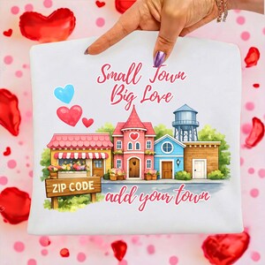 Puede incluir: Cuadrado decorativo blanco con una colorida ilustración de una escena de pueblo pequeño. El diseño incluye edificios, corazones y el texto "Small Town Big Love" y "add your town". Un letrero dice "ZIP CODE".