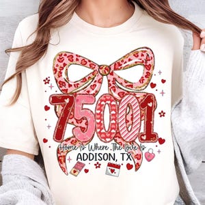 Puede incluir: Camiseta color crema con un gran lazo con diseño de estampado de leopardo y detalles de purpurina dorada. El número "75001" se muestra en rojo y rosa, con el texto "Home Is Where The Love Is ADDISON, TX". Corazones y detalles florales completan el diseño.