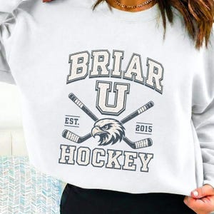 May include: A white crewneck sweatshirt featuring a dark blue graphic print. The design includes the text BRIAR U HOCKEY, EST. 2015, and an eagle head illustration positioned between two crossed hockey sticks in a distressed, vintage style.