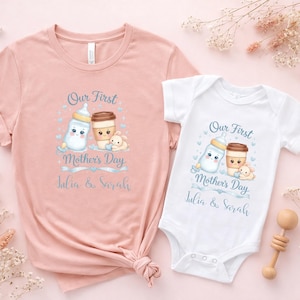 May include: A pink t-shirt and a white baby onesie with matching graphics. The design includes a baby bottle, coffee cup, and a teddy bear, with the text "Our First Mother's Day" and names. A wooden rattle is also visible.
