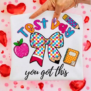 Puede incluir: Camiseta blanca con gráficos coloridos para un día de examen. El diseño incluye las palabras "Test Day" en azul, naranja y rosa, un lazo, una manzana, un lápiz, un cuaderno y la frase "you got this".