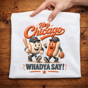 May include: White t-shirt featuring a baseball and hot dog cartoon characters wearing baseball caps. The shirt has the text "Hey Chicago" in orange and "WHADYA SAY!" in gray.