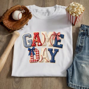 Könnte beinhalten: Weißes T-Shirt mit dem Schriftzug "GAME DAY" in einem Baseball-Design. Die Buchstaben sind mit Baseballnähten, Karomustern und Schleifen verziert. Ein Baseballhandschuh, ein Schläger, ein Ball, Popcorn und Jeansshorts sind ebenfalls im Bild.
