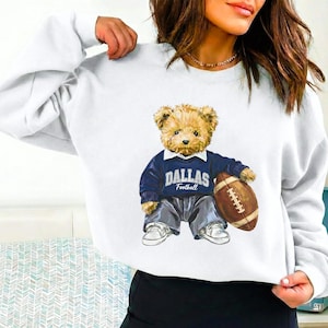 May include: White sweatshirt featuring a teddy bear wearing a navy blue sweater with "DALLAS Football" text and holding a brown football. The bear is also wearing gray pants and white sneakers.