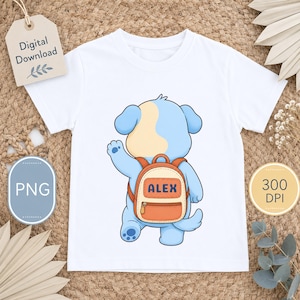 Op de afbeelding: Een wit t-shirt met een cartoonillustratie van een lichtblauwe puppy met een crèmekleurige vlek op zijn kop. De puppy is van achteren te zien en draagt een oranje rugzak met de naam ALEX in donkerblauwe letters erop gedrukt.