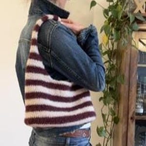 May include: A striped shoulder bag in burgundy and cream is carried over the shoulder. The bag's handle and body feature horizontal stripes. The bag is made of a soft, knitted material. The bag is worn by a person wearing a denim jacket.