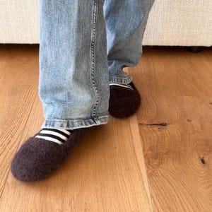 Könnte beinhalten: Ein Paar dunkelbraune Filzhausschuhe aus Wolle mit abgerundeter Zehenpartie. Die Hausschuhe weisen am oberen Rand ein sichtbares schwarz-weißes Querstreifenmuster auf. Das Schuhwerk wird mit einer hellblauen Jeans kombiniert, die über die Knöchel fällt.