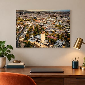 Peut inclure: Une impression murale rectangulaire présentant une vue aérienne d'une ville avec une haute tour d'horloge au premier plan, entourée de bâtiments résidentiels, d'arbres et de routes sous un ciel clair, exposée sur un mur au-dessus d'un bureau.