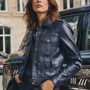 May include: A dark blue denim jacket with a classic design, featuring button closures and pockets. The jacket is worn over a dark top. The image is set outdoors with a car in the background.