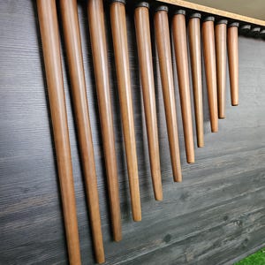 May include: A collection of wooden furniture legs, of varying lengths, are displayed in a row. The legs are a warm brown hue and have a tapered design. They are attached to a dark grey wooden surface. The background includes a strip of green artificial turf.