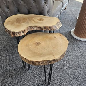 Puede incluir: Dos mesas auxiliares de madera con bordes naturales y patas de horquilla negras. Las mesas están hechas de madera de color claro y tienen una forma circular y orgánica. Las mesas están colocadas sobre una alfombra gris.