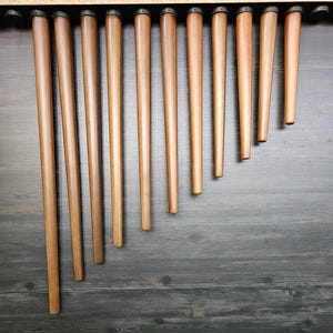 Puede incluir: Una colección de patas de muebles de madera en varios tonos de marrón, dispuestas en orden descendente. Cada pata tiene un diseño cónico, con una parte superior más ancha y una base más estrecha. Las patas están sobre un fondo de madera gris oscuro.