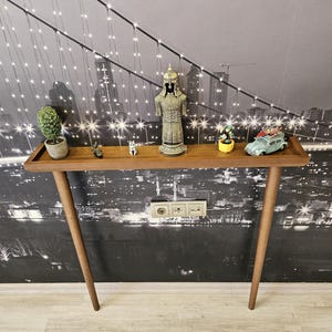 May include: A mid-century modern style wooden console table with a rectangular top and angled legs. The table displays decorative items, including a small statue, miniature car, and potted plants. The background features a black and white cityscape.