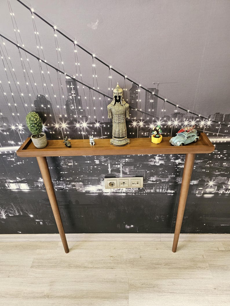 May include: A mid-century modern style wooden console table with a rectangular top and angled legs. The table displays decorative items, including a small statue, miniature car, and potted plants. The background features a black and white cityscape.