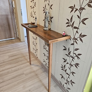 Op de afbeelding: Een houten console met een rechthoekig blad en vier slanke, taps toelopende poten. De tafel heeft een warme bruine kleur en een klein plankje. Decoratieve items, waaronder een beeldje en een kleine fles, worden op het tafelblad getoond.