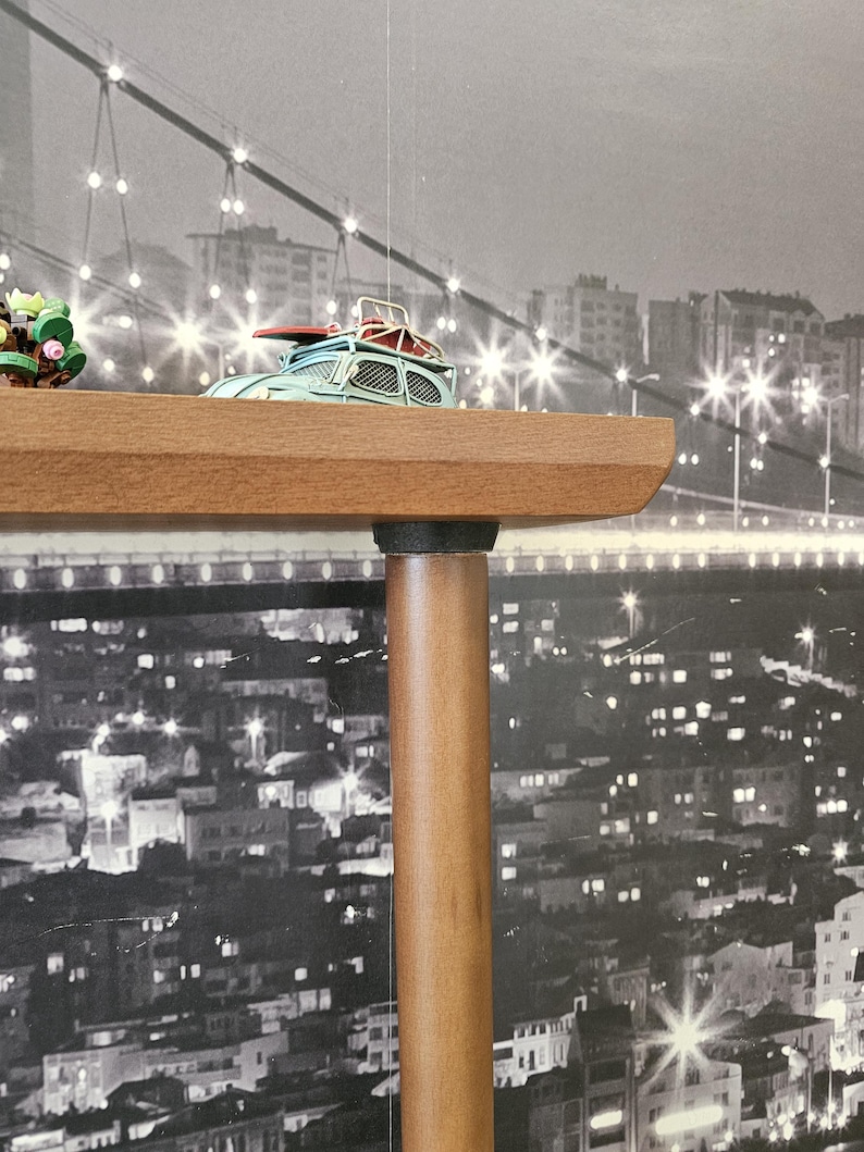 May include: A light blue toy car with a red surfboard on top sits on a wooden shelf. The shelf is supported by a brown wooden leg. The background is a black and white cityscape with a bridge.