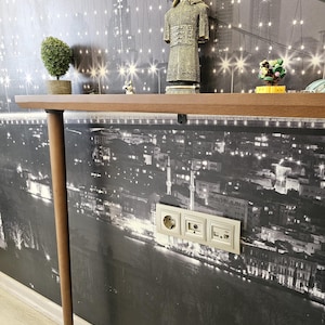 May include: A brown wooden shelf with a single leg, displaying decorative items against a black and white cityscape wallpaper. Items include a small topiary, a figurine of a warrior, and other small ornaments. Electrical outlets are visible on the wall.