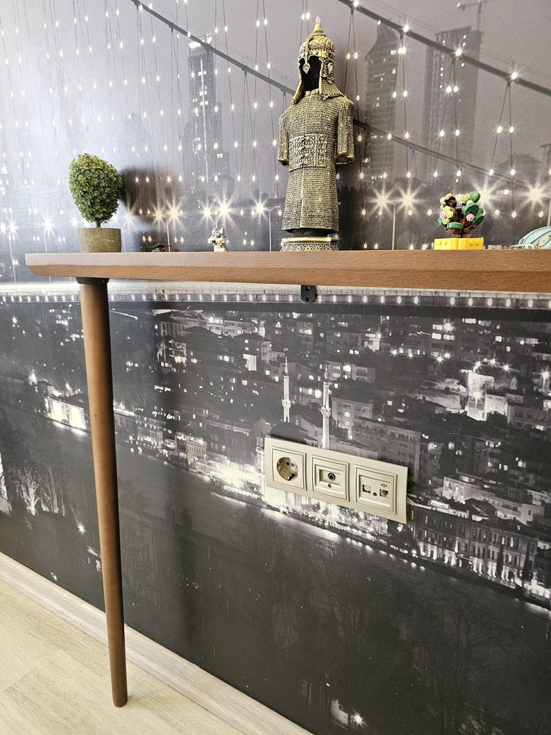 May include: A brown wooden shelf with a single leg, displaying decorative items against a black and white cityscape wallpaper. Items include a small topiary, a figurine of a warrior, and other small ornaments. Electrical outlets are visible on the wall.