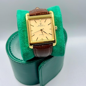 May include: A gold-toned Rolex Oyster Perpetual Date automatic watch with a rectangular face and brown leather band. The watch is displayed on a green velvet cushion, with a green leather watch case in the background.