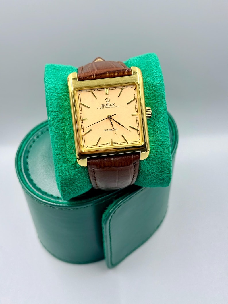 May include: A gold-toned Rolex Oyster Perpetual Date automatic watch with a rectangular face and brown leather band. The watch is displayed on a green velvet cushion, with a green leather watch case in the background.