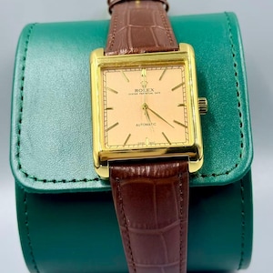 May include: A gold-toned Rolex Oyster Perpetual Date automatic watch with a rectangular face and brown leather band. The watch has a light peach-colored dial with gold hands and markers. The watch is displayed on a green leather case.