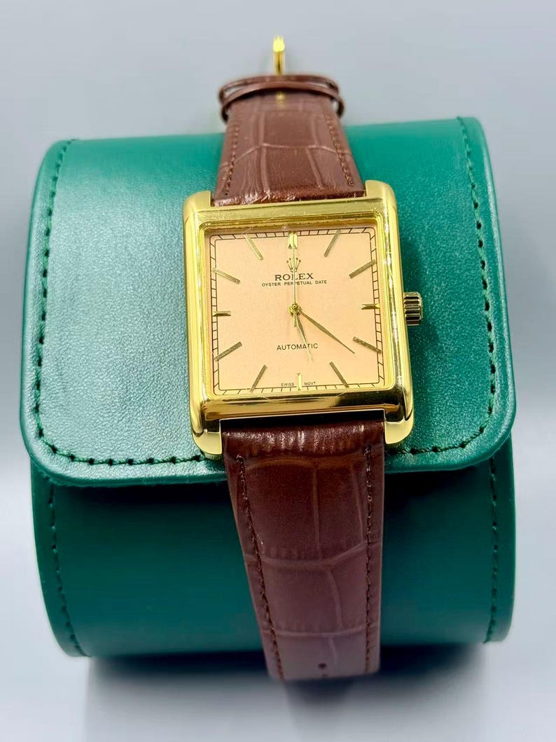 May include: A gold-toned Rolex Oyster Perpetual Date automatic watch with a rectangular face and brown leather band. The watch has a light peach-colored dial with gold hands and markers. The watch is displayed on a green leather case.