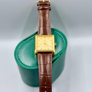 May include: A gold rectangular wristwatch with a brown leather band. The watch has a gold face with black hands and markers. The watch is resting on a green leather case. The band is approximately 0.75 inches wide.