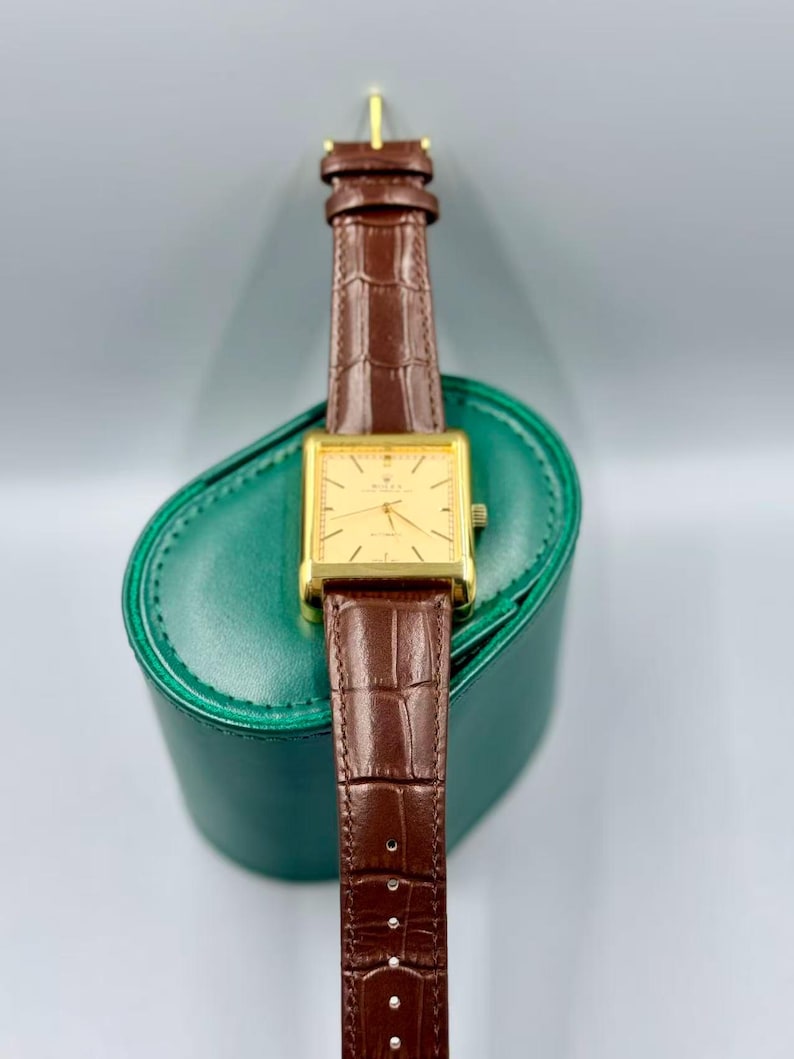 May include: A gold rectangular wristwatch with a brown leather band. The watch has a gold face with black hands and markers. The watch is resting on a green leather case. The band is approximately 0.75 inches wide.
