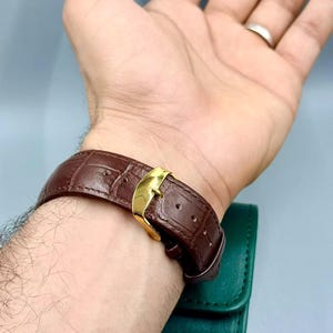 May include: A brown leather watch band with a gold-colored buckle is fastened on a wrist. The band has a textured, crocodile-like pattern. A green watch case is visible in the background.
