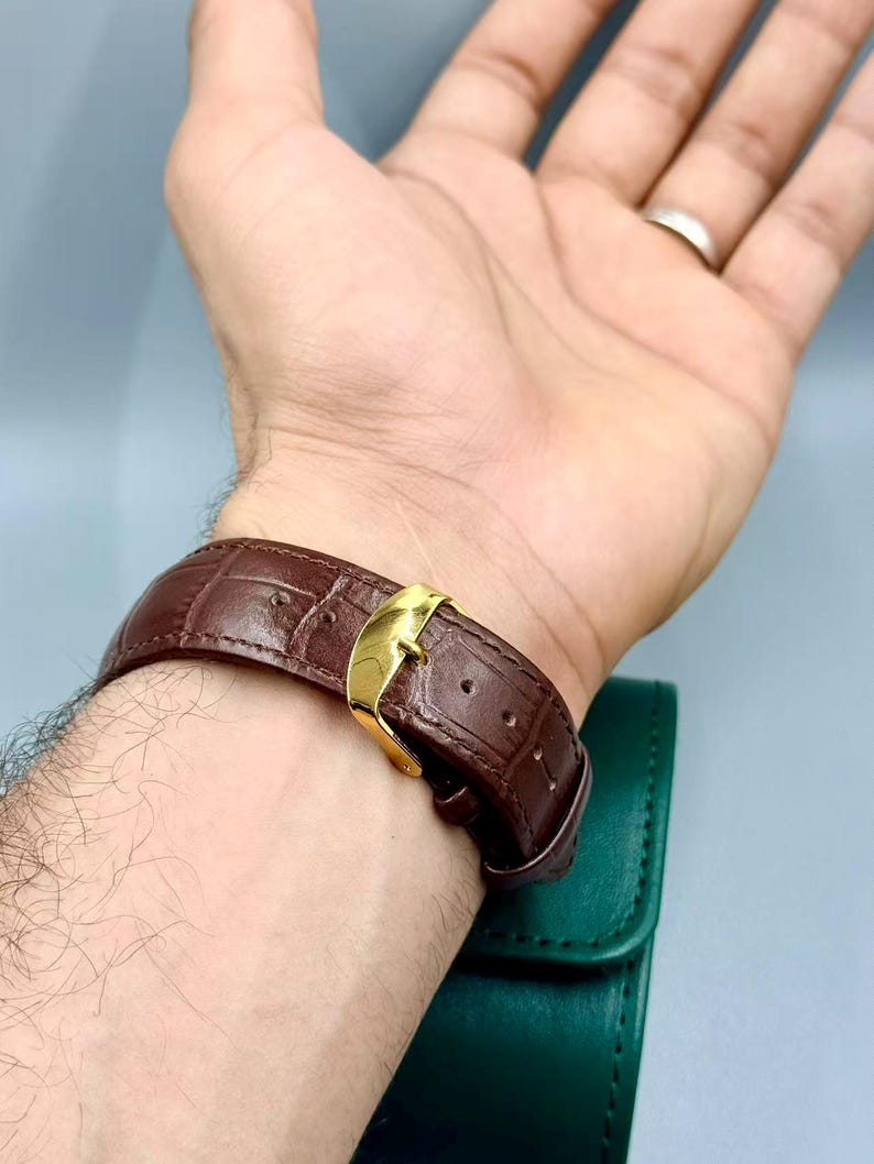 May include: A brown leather watch band with a gold-colored buckle is fastened on a wrist. The band has a textured, crocodile-like pattern. A green watch case is visible in the background.
