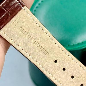 May include: Close-up of a brown leather watch strap with the text "GENUINE LEATHER" stamped on the underside. The strap has a textured surface and is stitched with white thread. The watch strap is next to a green case.