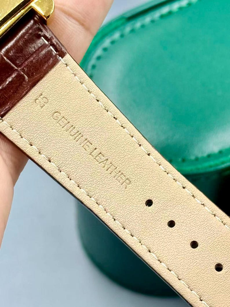 May include: Close-up of a brown leather watch strap with the text "GENUINE LEATHER" stamped on the underside. The strap has a textured surface and is stitched with white thread. The watch strap is next to a green case.