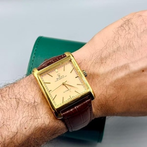 May include: A gold-toned rectangular Rolex Oyster Perpetual Date automatic watch with a light-colored face and brown leather band. The watch has a gold bezel and a brown leather strap. The watch is on a wrist.